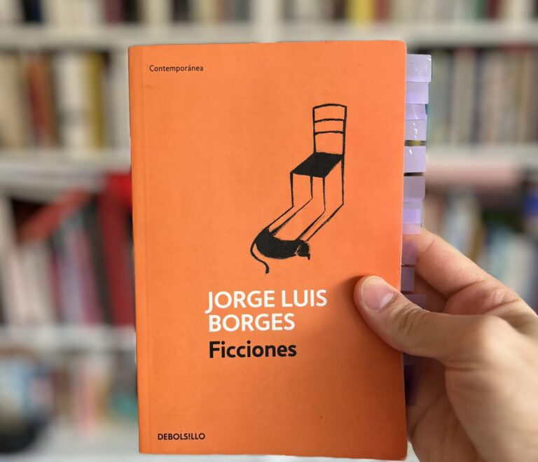 Libro "Ficciones" de Jorge Luis Borges, con portada naranja y dibujo de una silla, sostenido en una mano, con estantería de libros desenfocada al fondo.